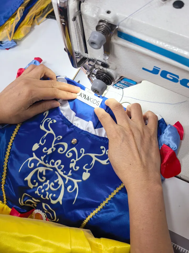 Twee handen stikken een Joya Kids-label in de hals van een blauwe prinsessenjurk