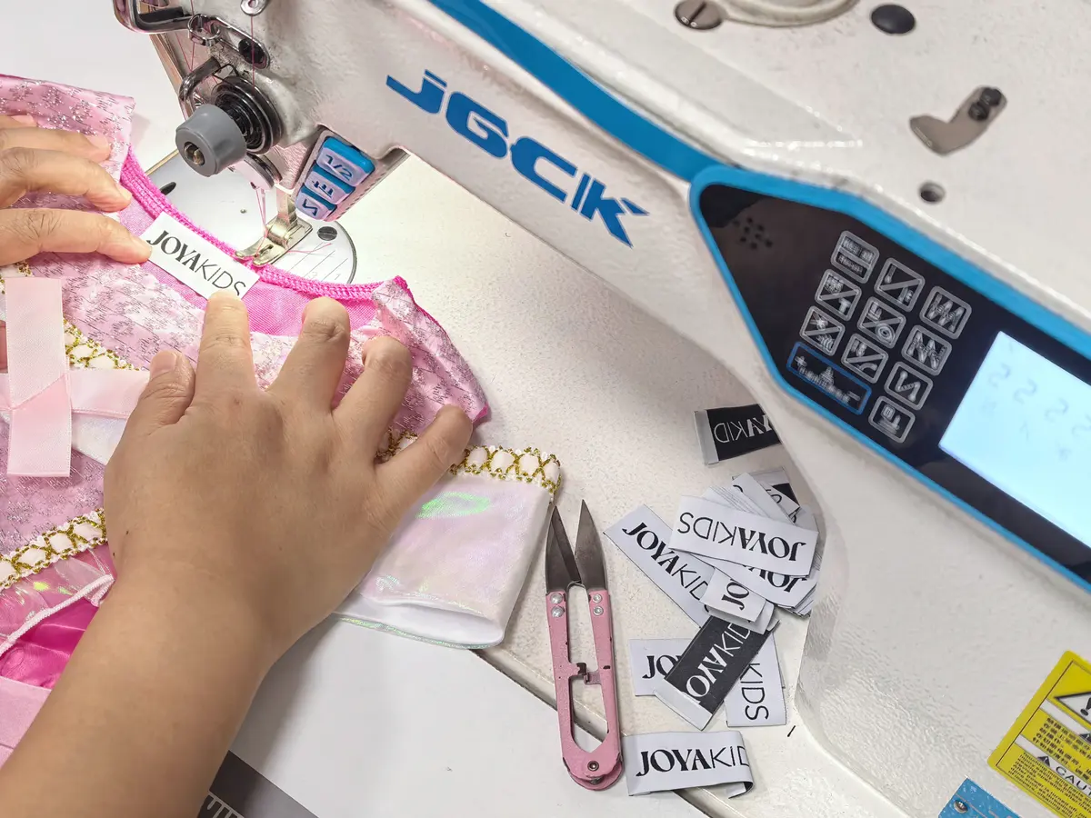 Twee handen stikken een Joya Kids-label in de hals van een roze verkleedjurk, met losse labels naast de naaimachine